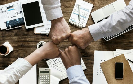 Four team members fist bumping to boost morale in the office above a table filled with papers and technology